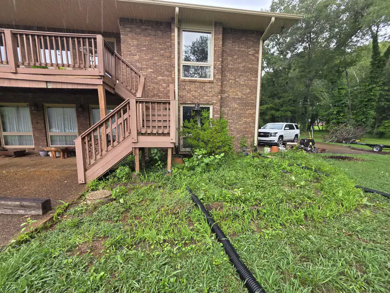 Weedy, unkempt ground under a raised deck with drain lines lying on the surface, before mulching.